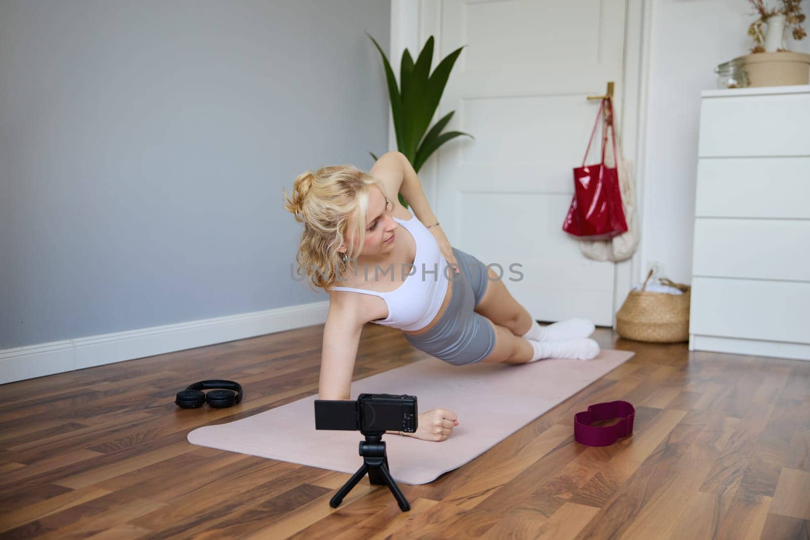 Portrait of young woman, fitness instructor recording video of her standing in a side plank, doing workout at home on camera, creating content for social media.