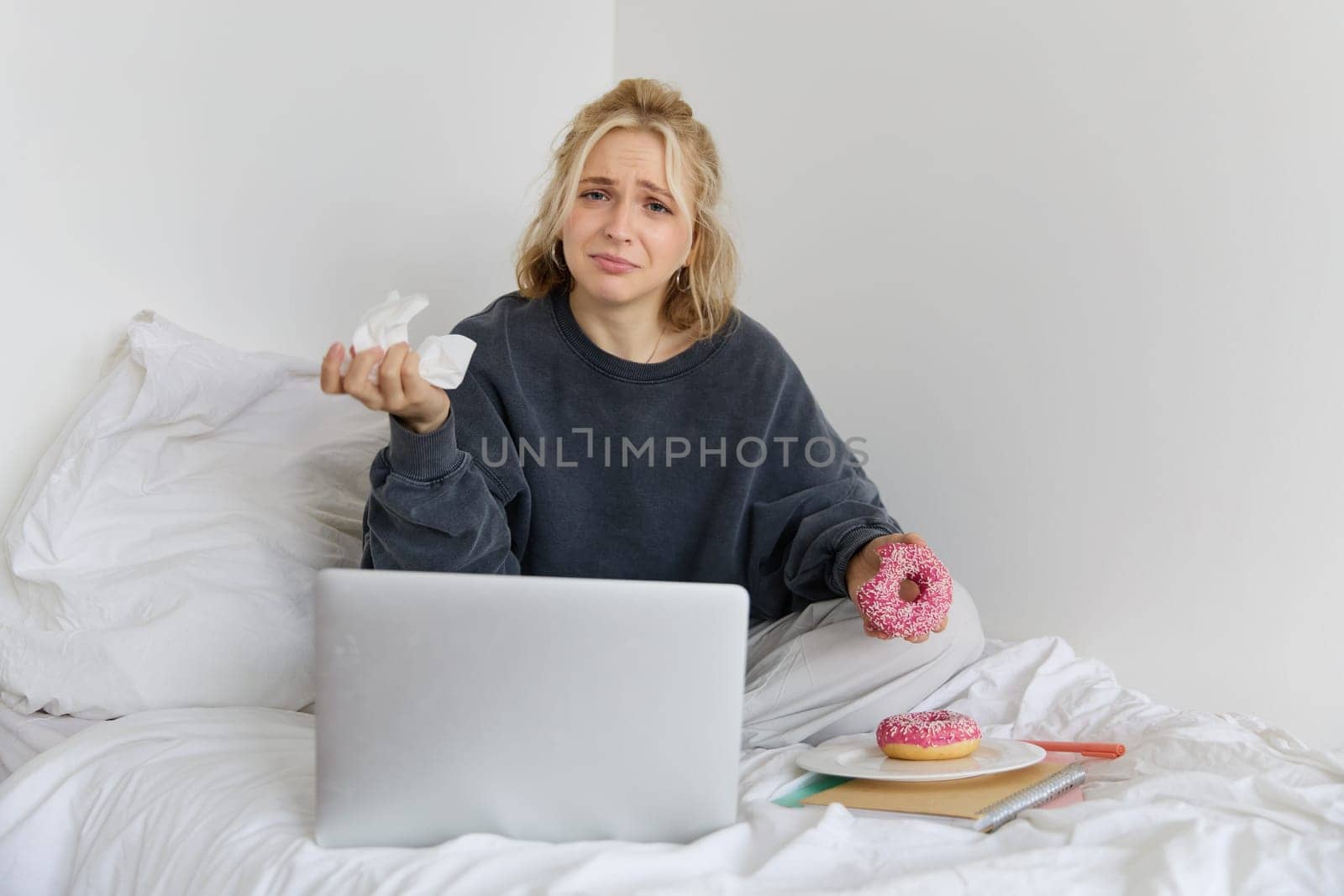 Portrait of disappointed, depressed young woman, crying, sitting on bed with laptop, eating comfort food, holding doughnut and wiping tears off.