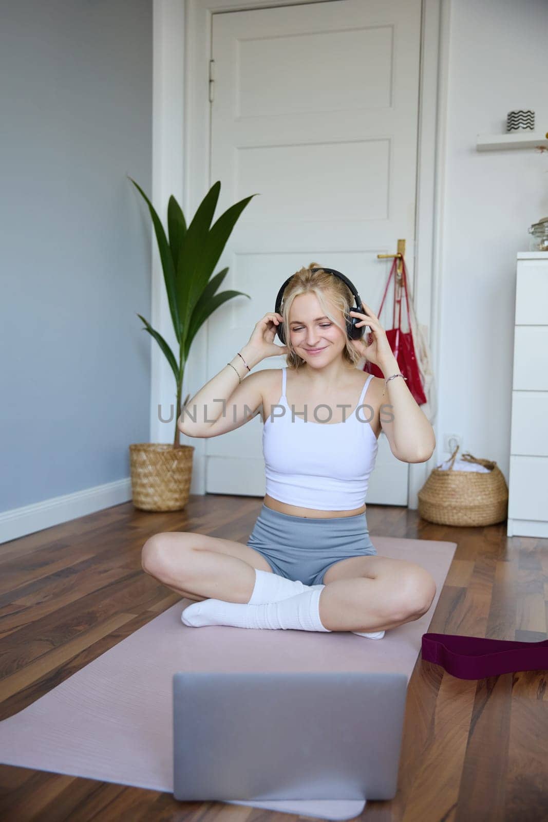 Portrait of young woman at home, connects to online gym training session, following fitness instructions on laptop, wearing wireless headphones during workout by Benzoix