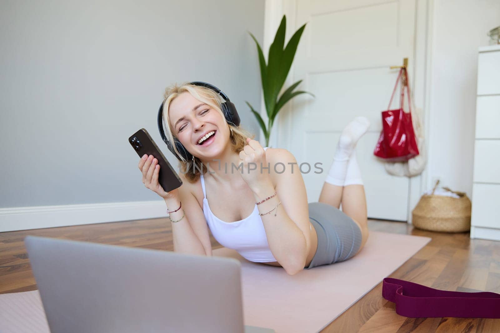 Portrait of woman lying on rubber yoga mat in room, wearing headphones, listens to music, using laptop and smartphone by Benzoix