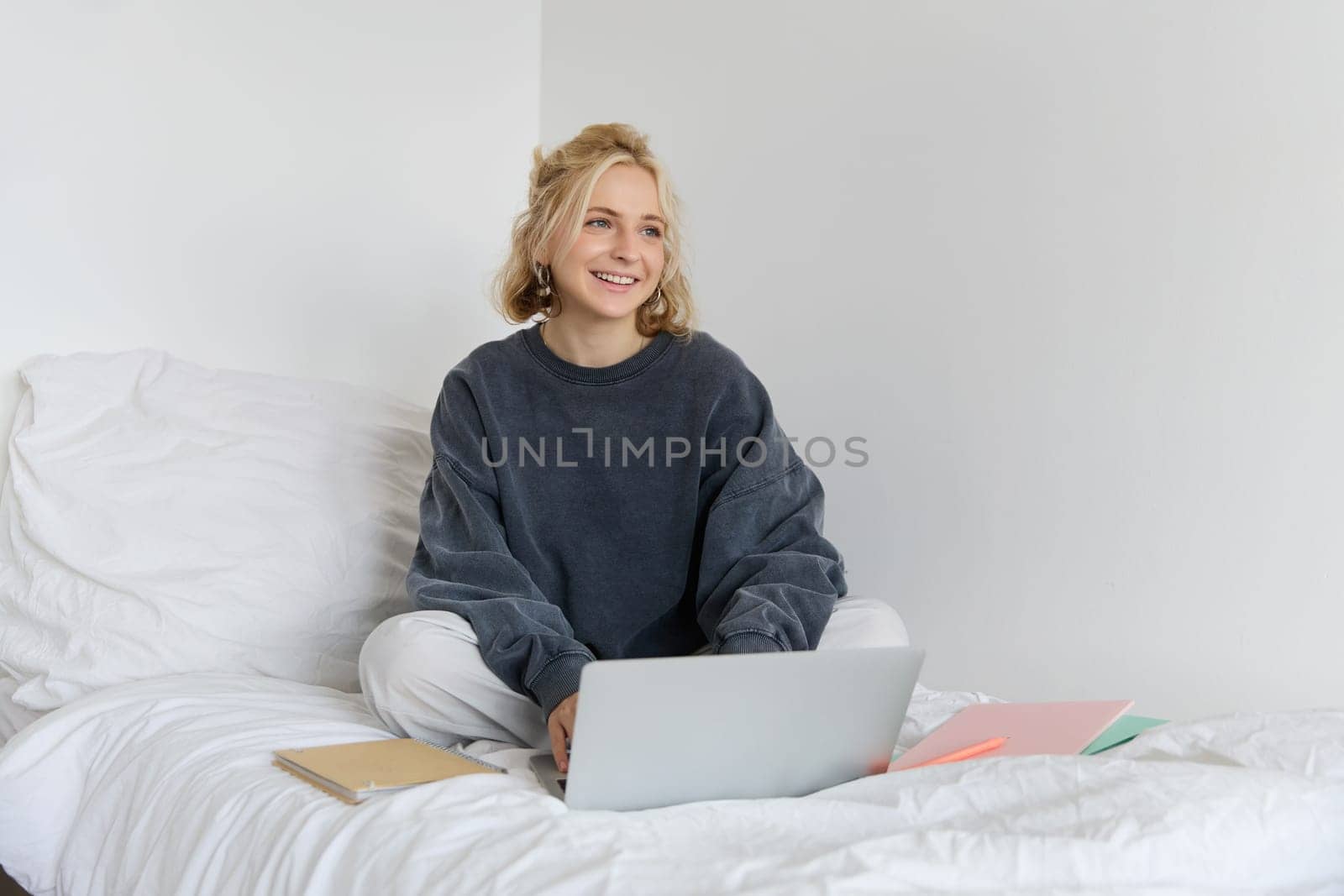 Image of happy young woman, student e-learning from home, connect to online course on her laptop, sits on bed with notebooks, smiling and looking happy.