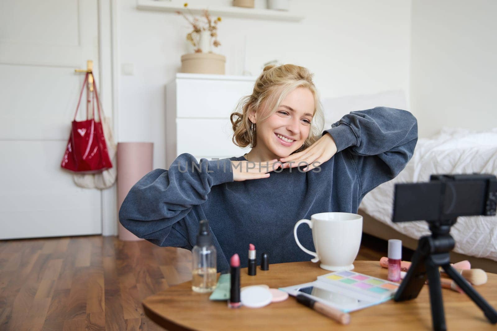 Lifestyle blogger records video of herself while doing hairstyle, showing makeup lifehacks for social media followers, vlogging, using camera for her blog, sitting in a room, drinking tea.