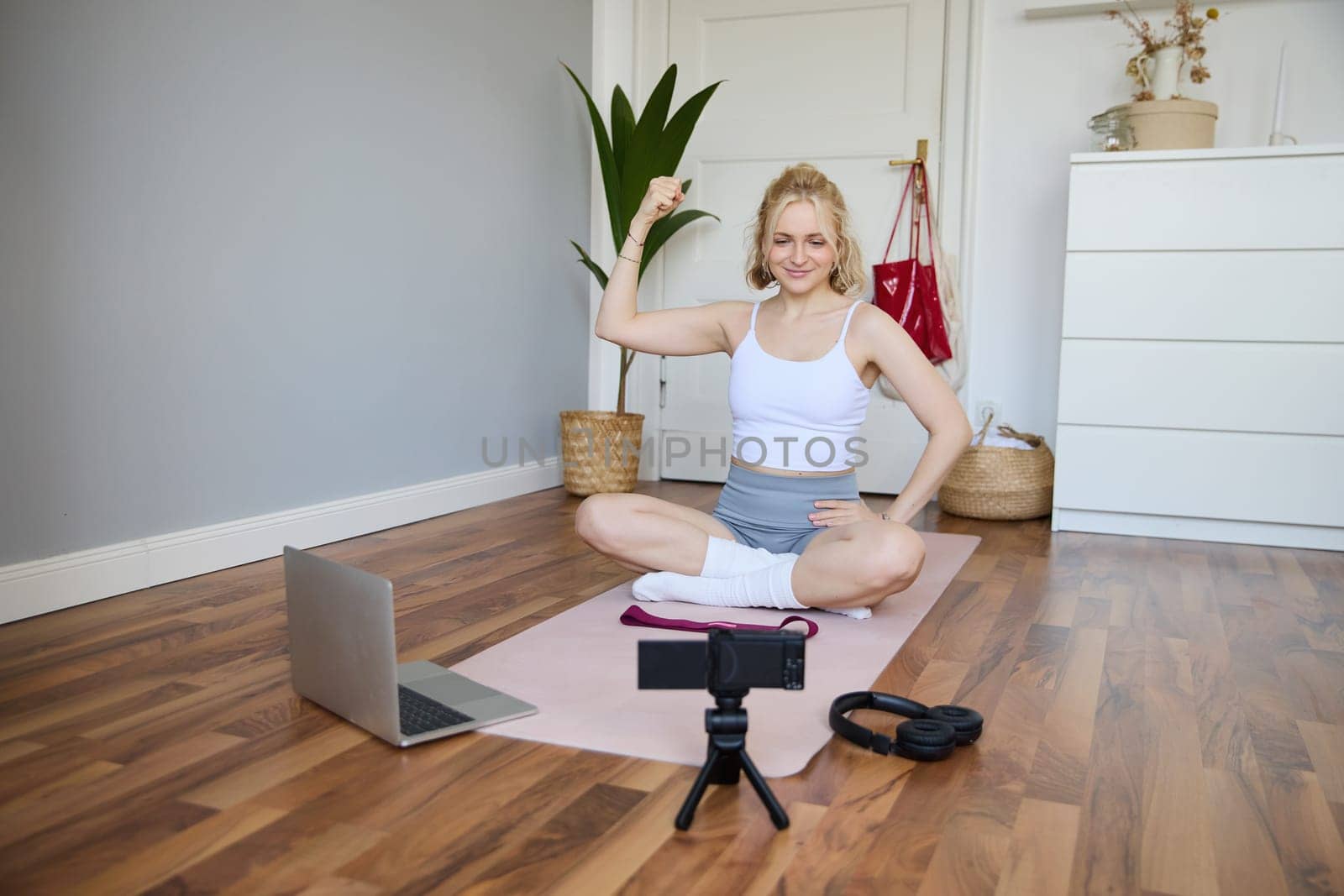 Portrait of young fitness instructor, vlogger showing exercises on camera, recording herself, sitting on mat with laptop, doing workout, explaining yoga movements to followers.