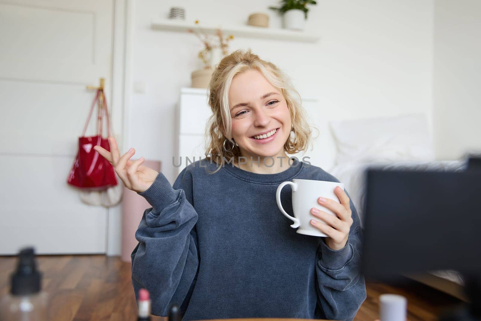 Portrait of cute smiling woman, blond girl drinks tea and records a casual, lifestyle video blog, vlogger sits in a room with camera and stabiliser, holds cup of coffee and talks.