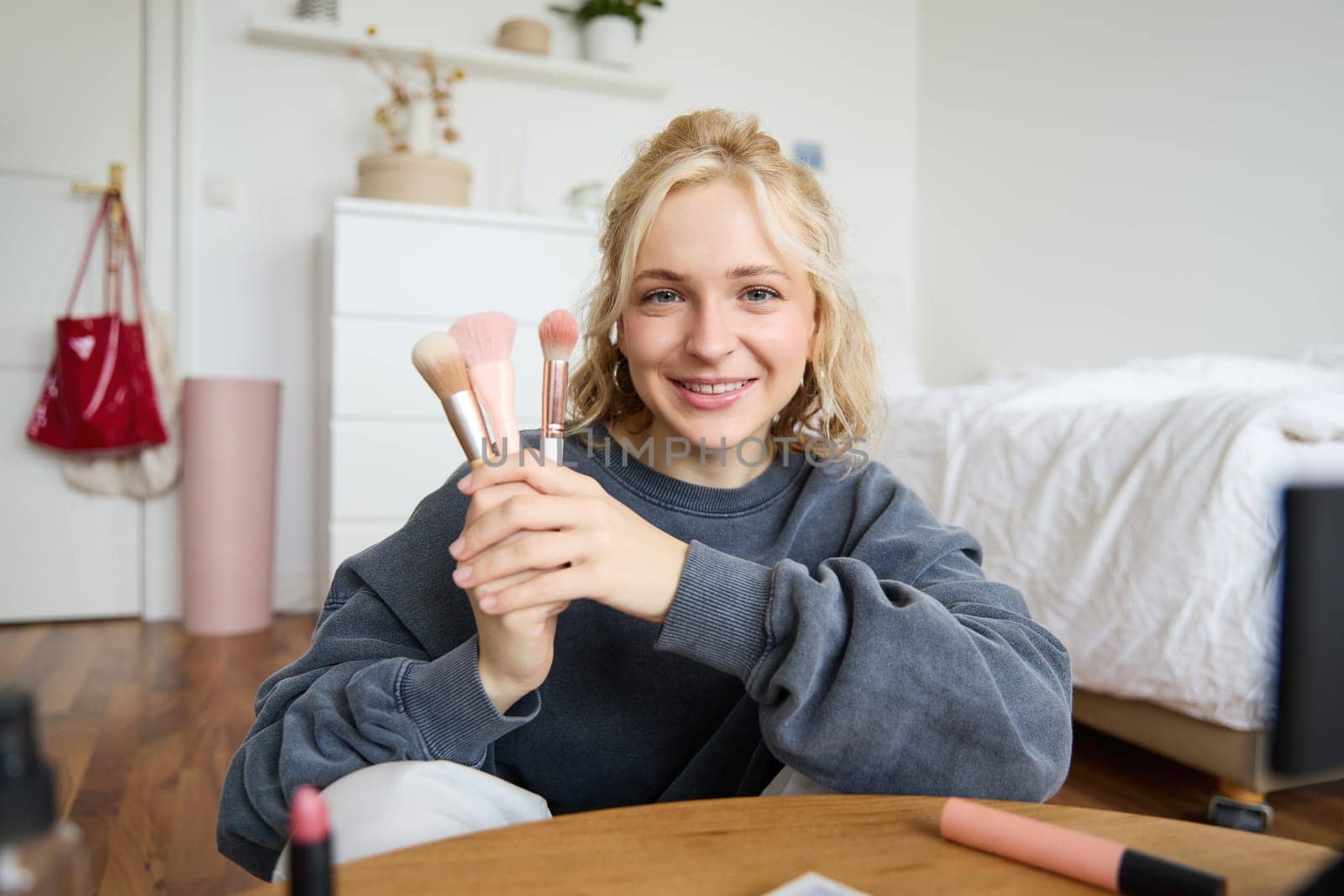 Portrait of young woman, content creator, making a video about makeup, showing brushes to audience, looking at camera, recording beauty tutorial, smiling happily.