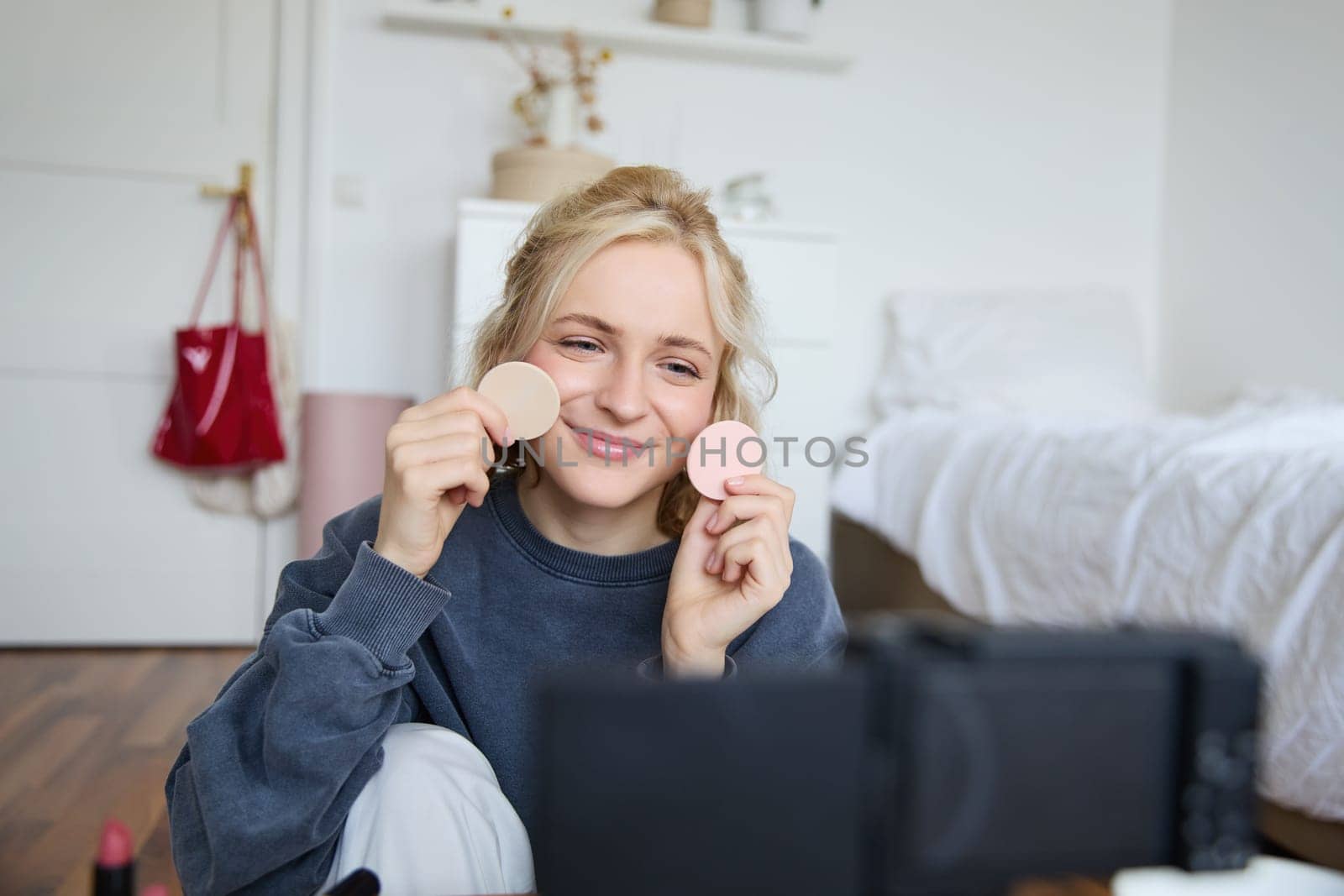 Portrait of young woman, beauty content creator, sitting in a room in front of digital camera, recording makeup tutorial vlog, showing cosmetic facial products.