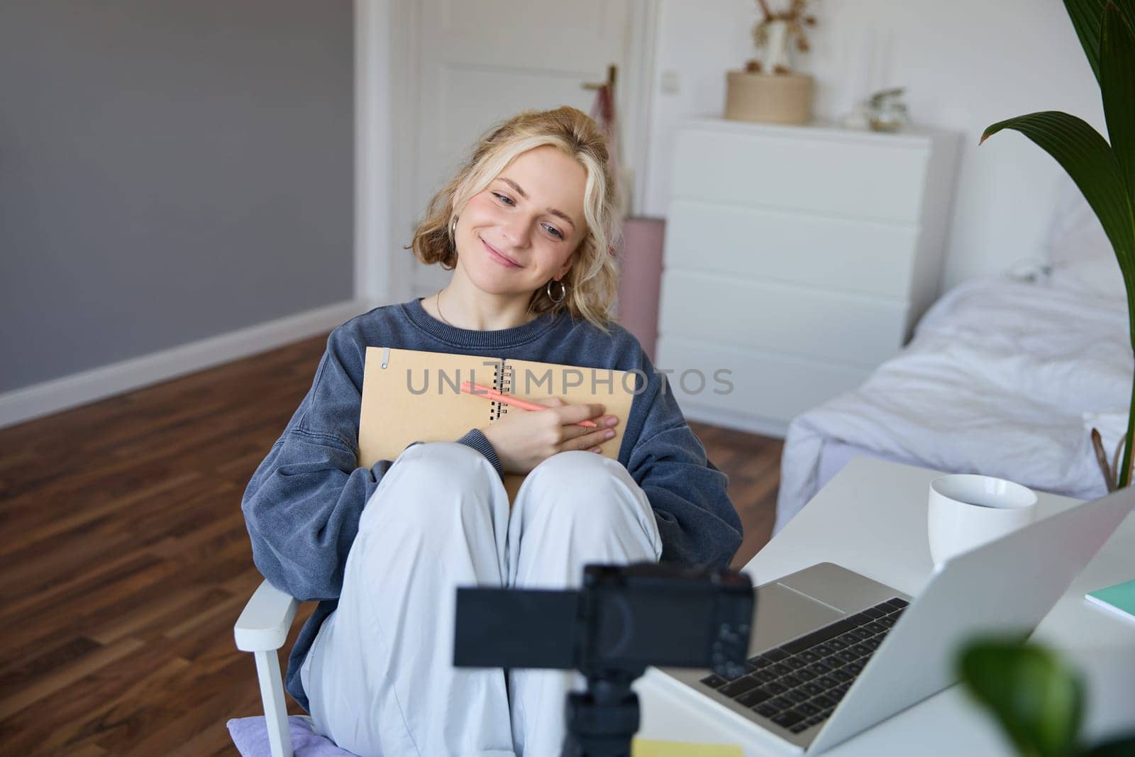 Portrait of blond smiling woman, records video on digital camera how she writes in notebook, talks to followers, doing lifestyle blog content in her room.