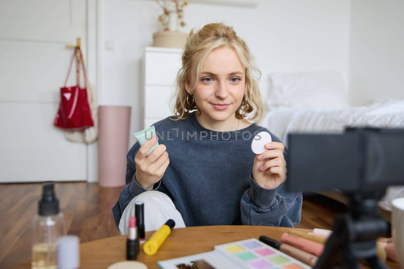 Portrait of young beauty blogger, woman gives advices to audience, how to apply makeup, sits in front of digital camera, holds foundation cream and cotton pad.
