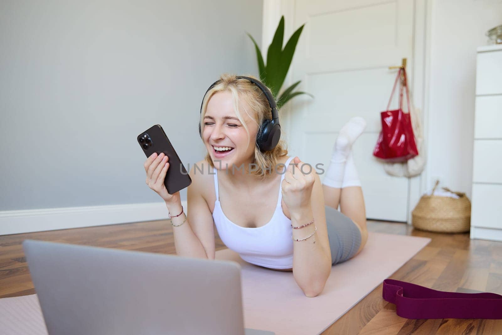 Portrait of young woman workout, watching exercise videos on laptop in headphones, lying on rubber mat with mobile phone and smiling by Benzoix