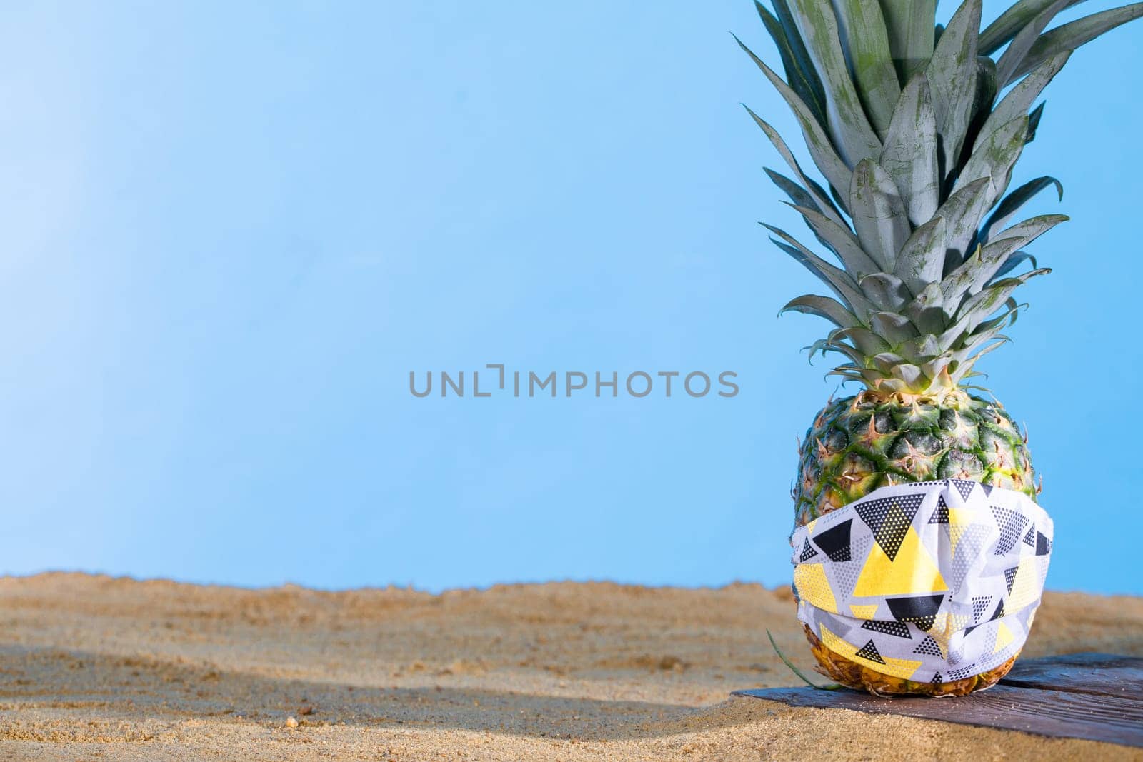 Pineapple in a protective mask stands on the sandy sea beach.