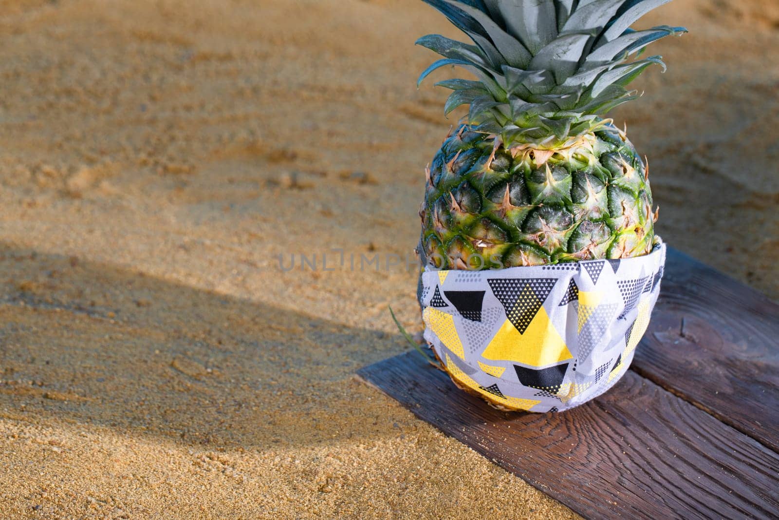 Pineapple in a protective mask stands on the sandy sea beach.