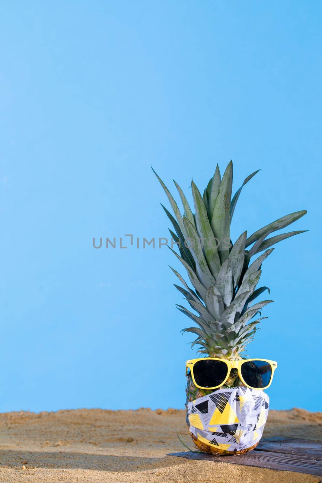 Pineapple in sun glasses and a protective mask stands on the sandy sea beach. by fotodrobik