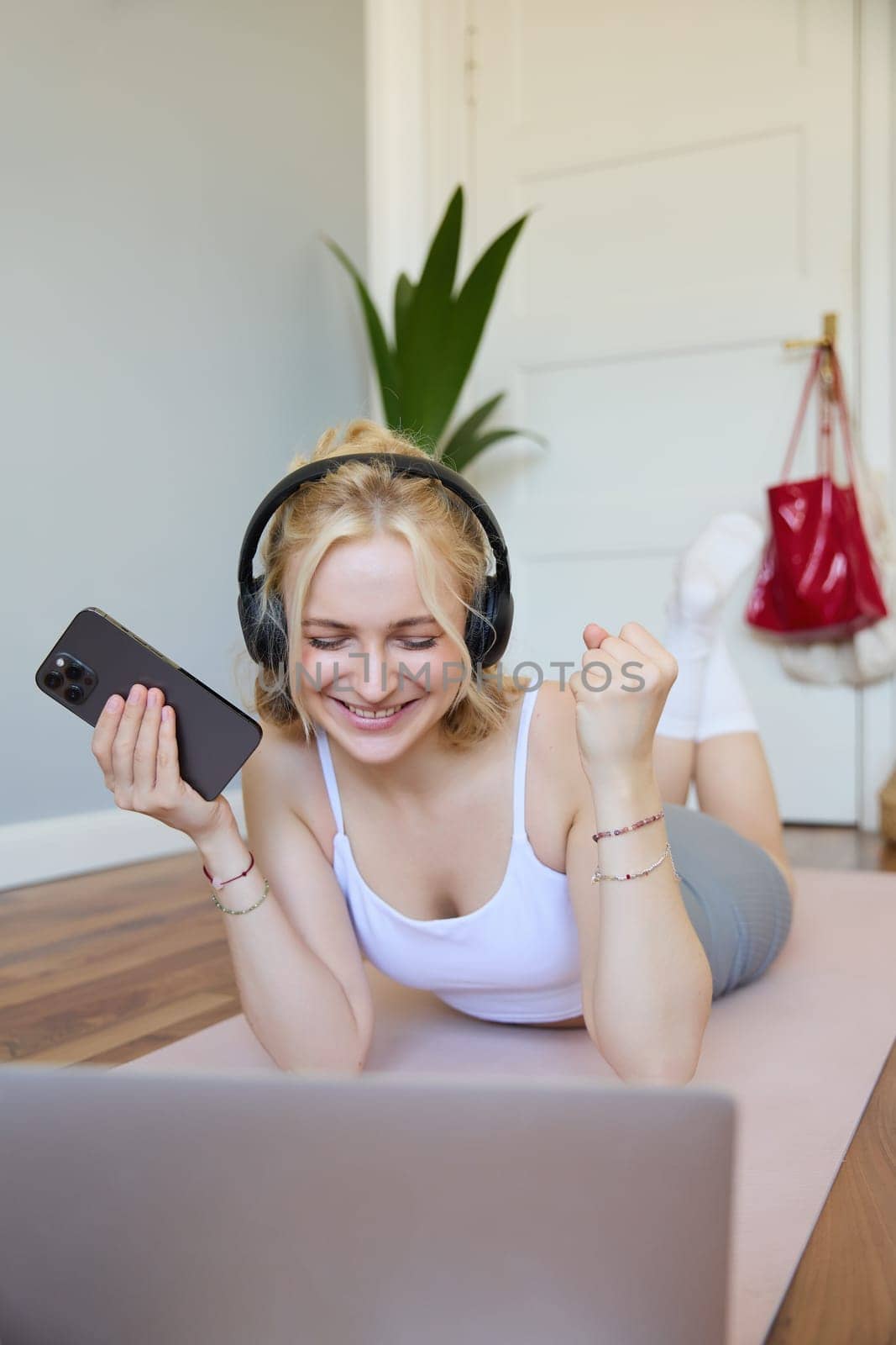 Vertical shot of young fitness woman in headphones, lying on rubber mat, relaxing after workout training session, holding smartphone, smiling at camera. Lifestyle and wellbeing concept