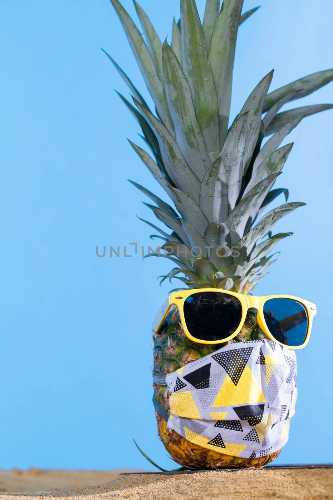 Pineapple in sun glasses and a protective mask stands on the sandy sea beach. by fotodrobik