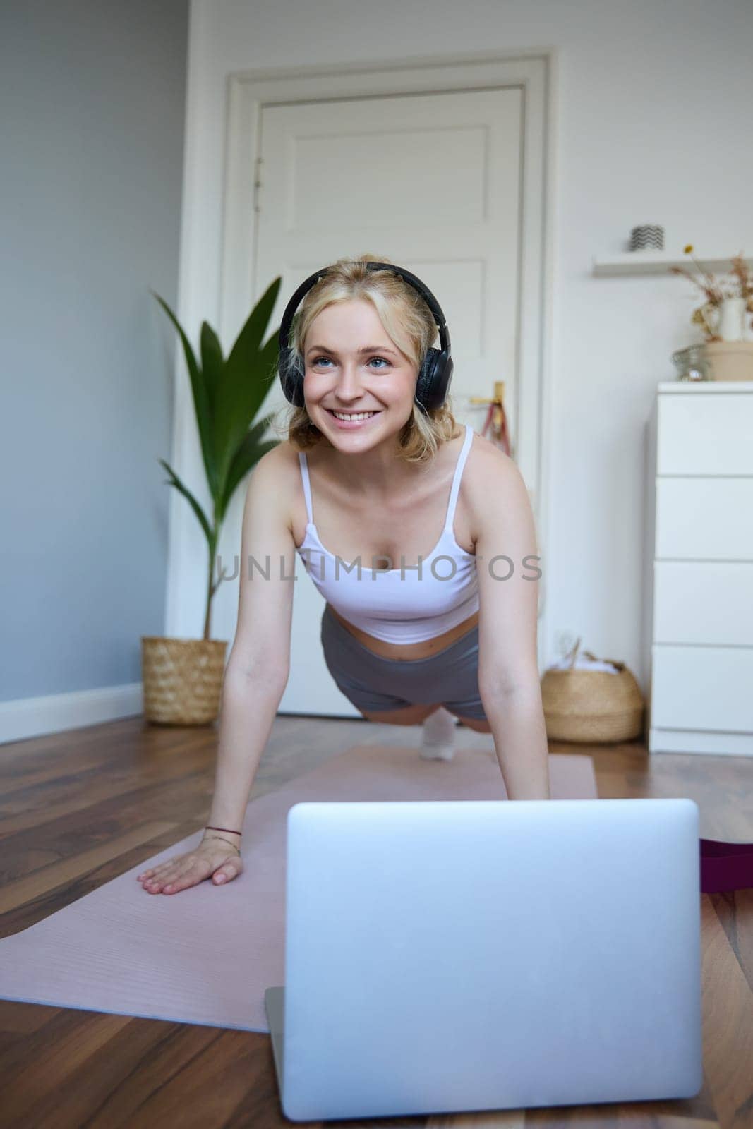 Portrait of young athletic woman in wireless headphones, standing in plank, following online workout video, using laptop to join training session on the internet.