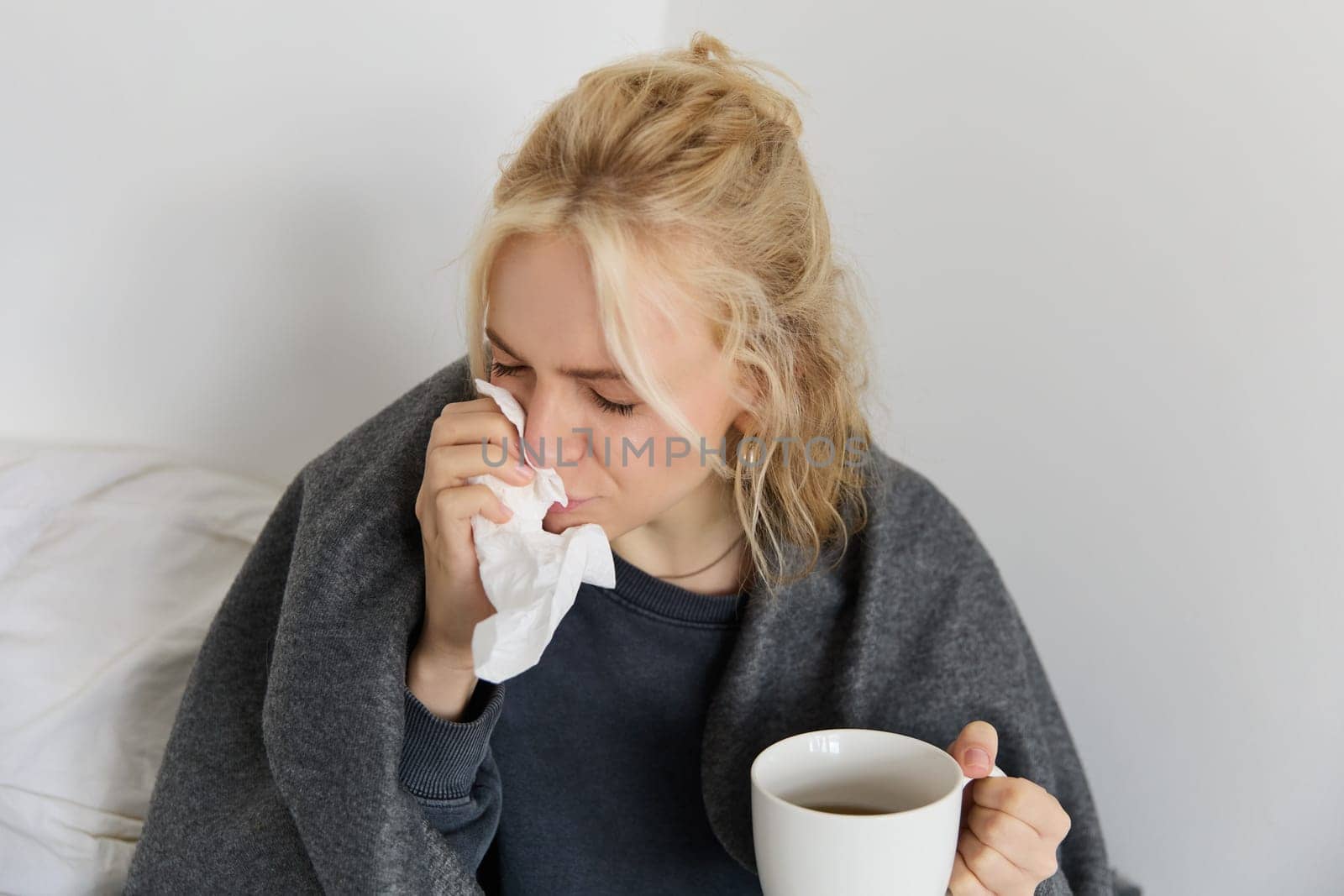 Concept of flu and quarantine. Close up portrait of young woman feeling sick, caught a cold, sneezing and blowing nose in napkin, holding warm tea in hand by Benzoix