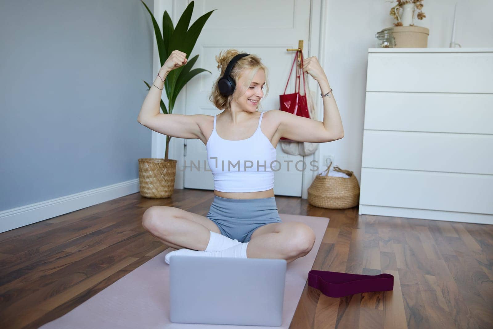 Portrait of young athletic woman doing workout at home, shows her muscles, strong biceps, sits on yoga mat and wears wireless headphones by Benzoix