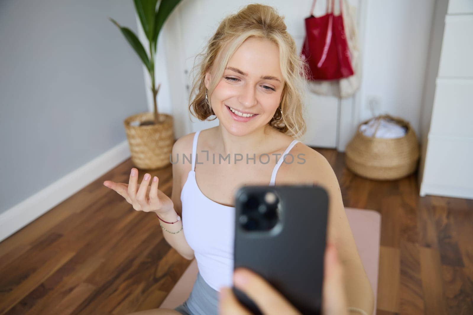 Portrait of beautiful young fitness trainer, showing home workout, taking selfie on mobile phone while sitting on yoga rubber mat in bedroom, wearing activewear.