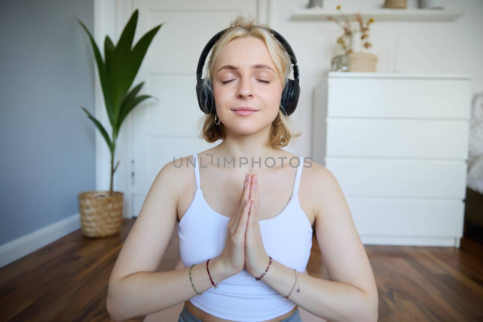 Portrait of woman meditating at home, sitting in headphones, listening to yoga podcast, holding hands in namaste sign.