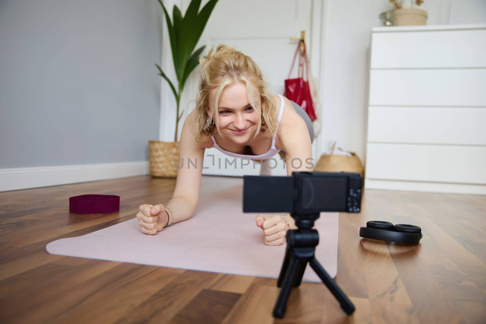 Portrait of young sporty vlogger, fitness instructor standing in plank on rubber yoga mat, recording video of herself doing workout at home.