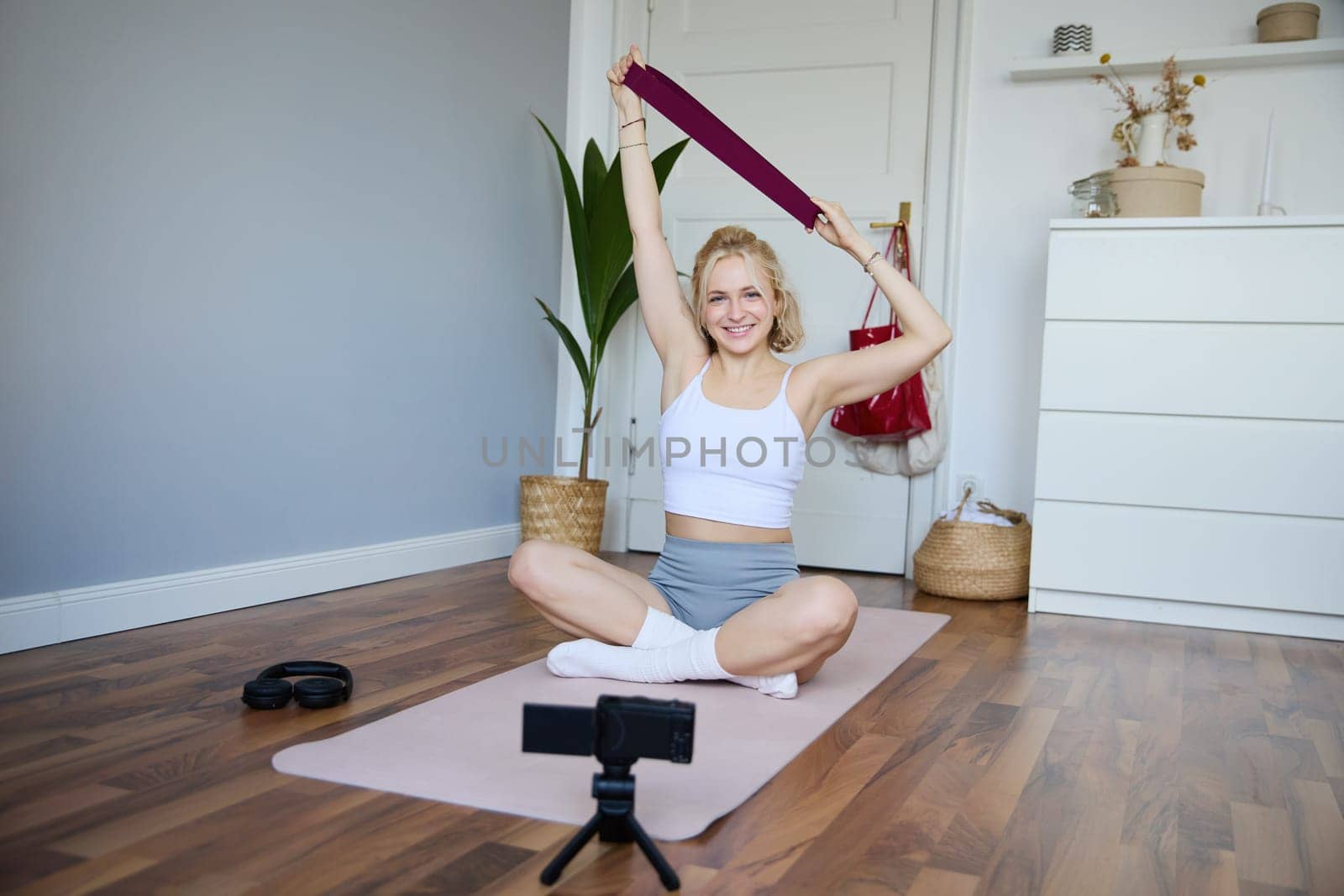 Image of smiling, athletic fitness instructor, female social media influencer, recording video about workout, showing how to use resistance bands on digital camera, sitting on rubber yoga mat at home.