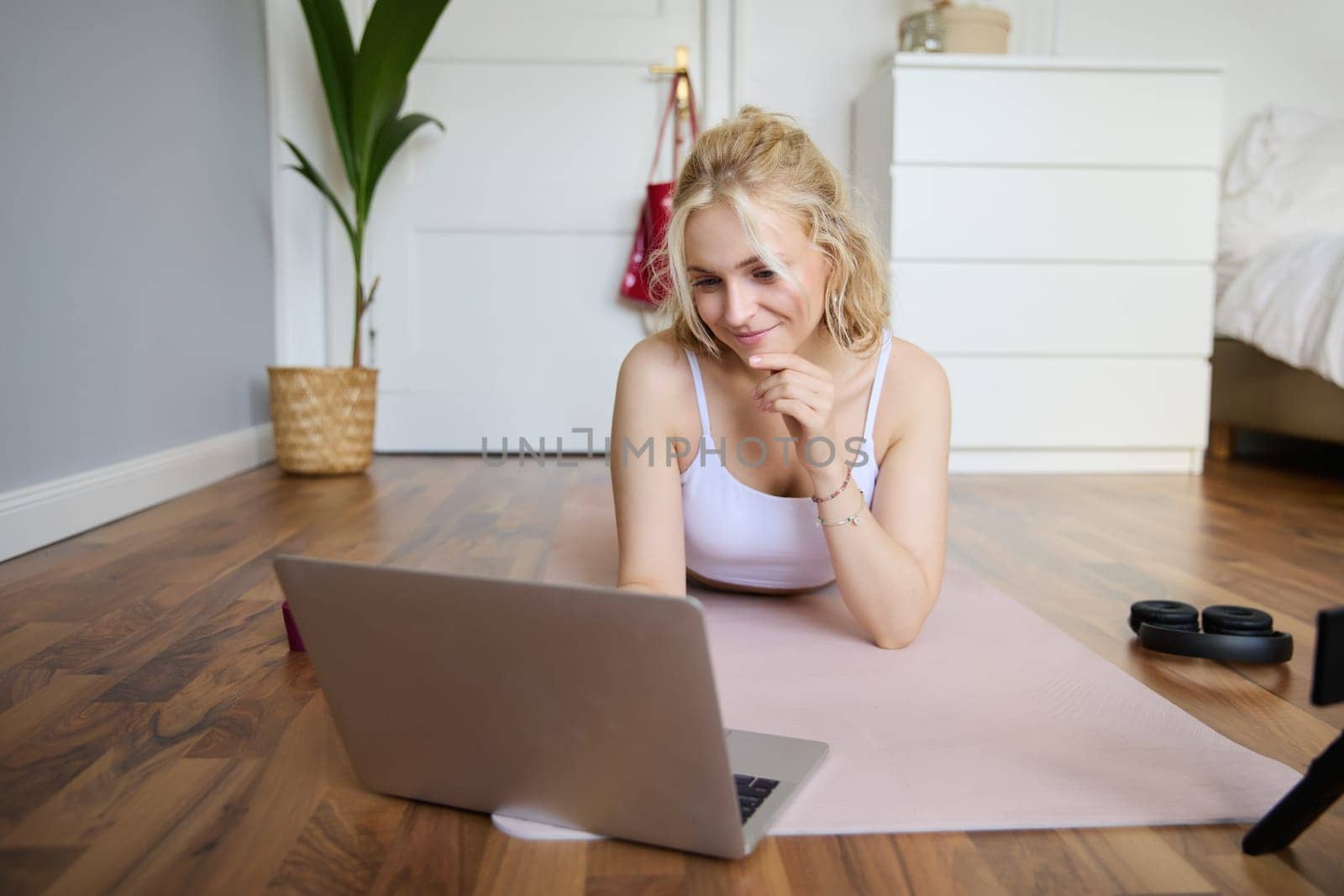Portrait of beautiful blond woman looking at fitness video tutorials on laptop, lying on rubber yoga mat, following workout instructions online by Benzoix