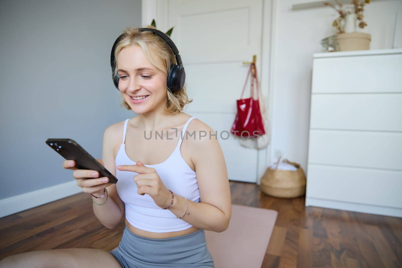 Portrait of young sporty woman in wireless headphones, sitting with smartphone on rubber mat, using workout training app, fitness application on her phone, listening in earphones by Benzoix