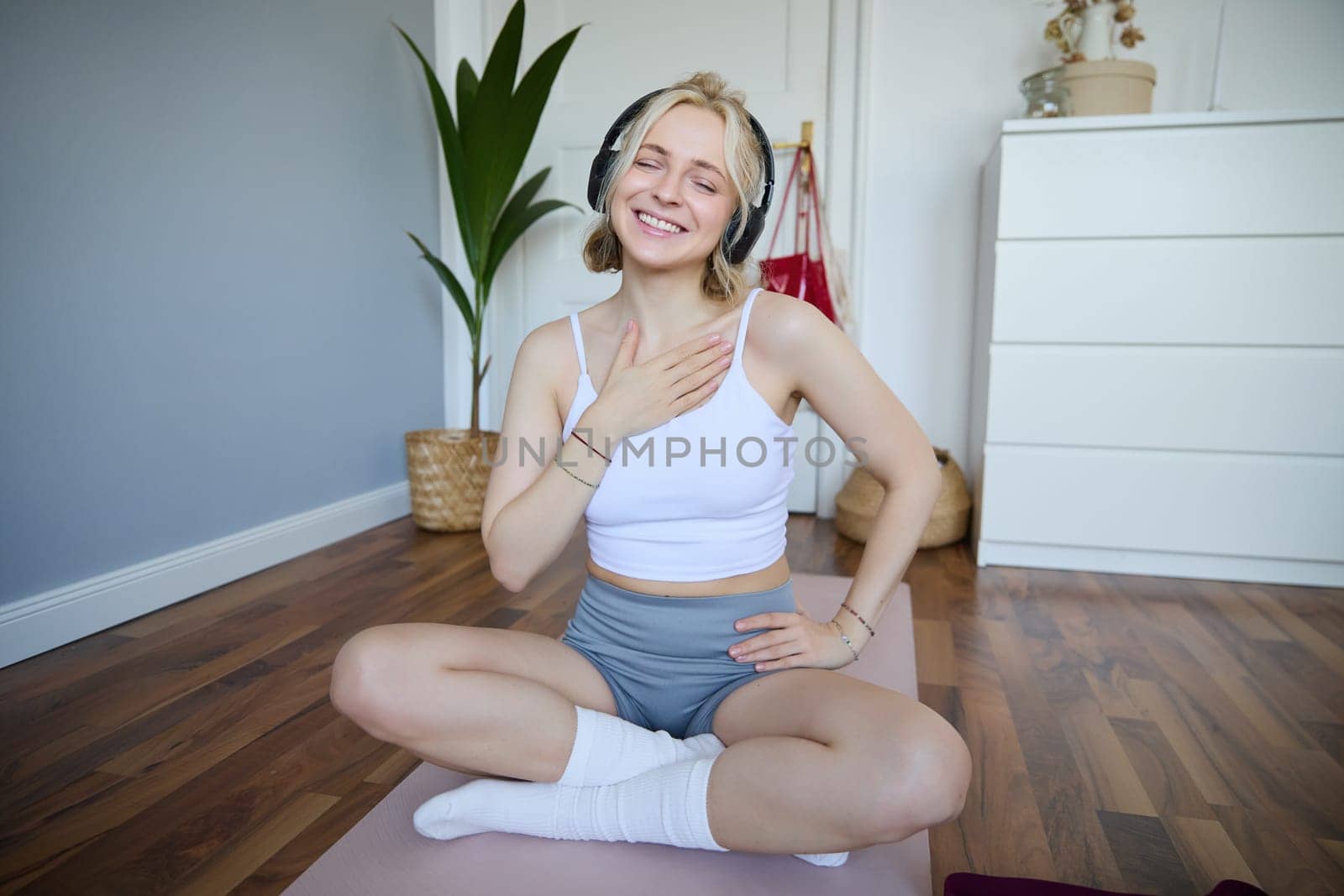 Portrait of young fitness woman, sporty female model in her room, sits on yoga mat, listens to music during workout, does exercises by Benzoix