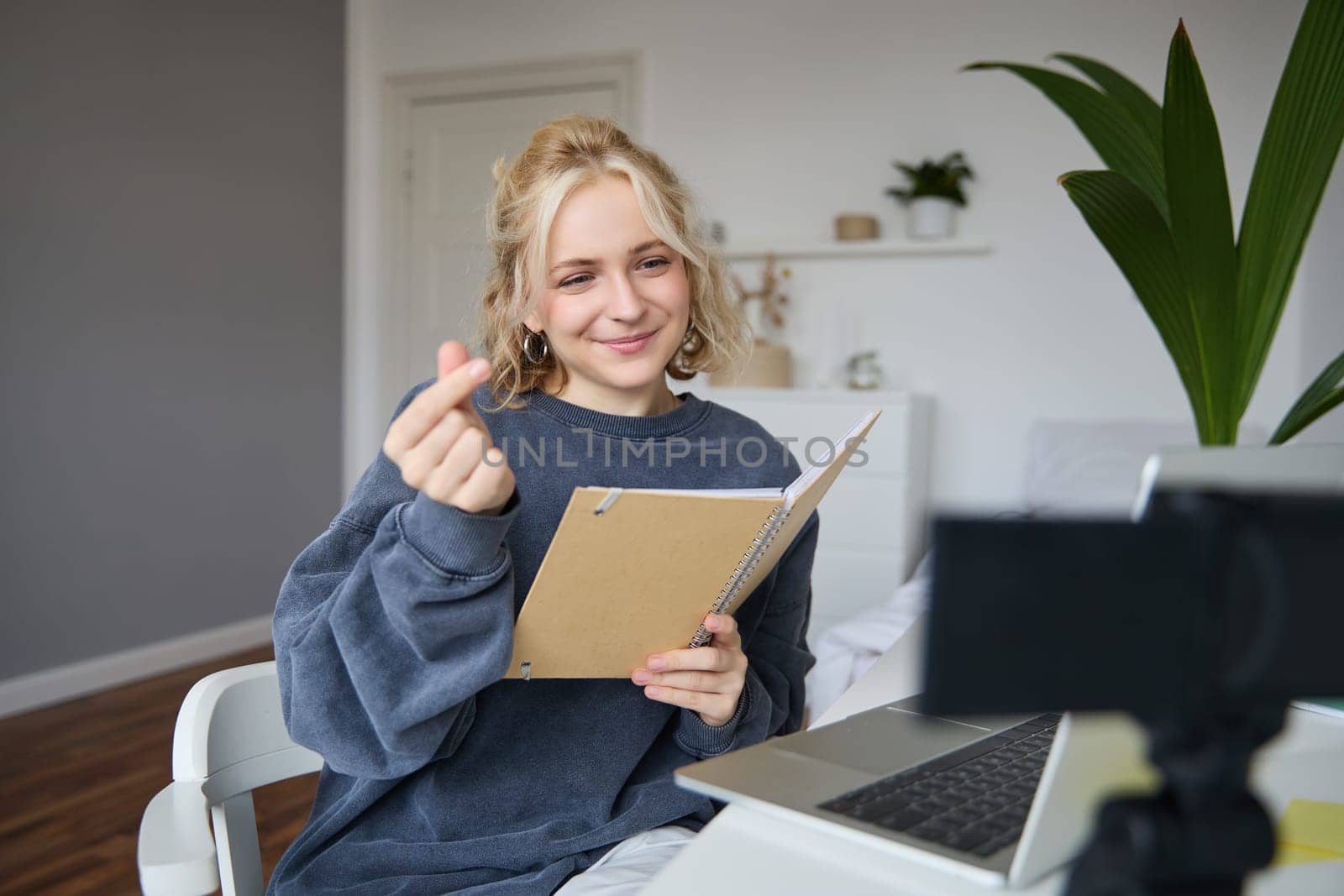 Beautiful young woman in her room, sits indoors, records video on digital camera, holds notebook and her notes, shows heart finger sign.