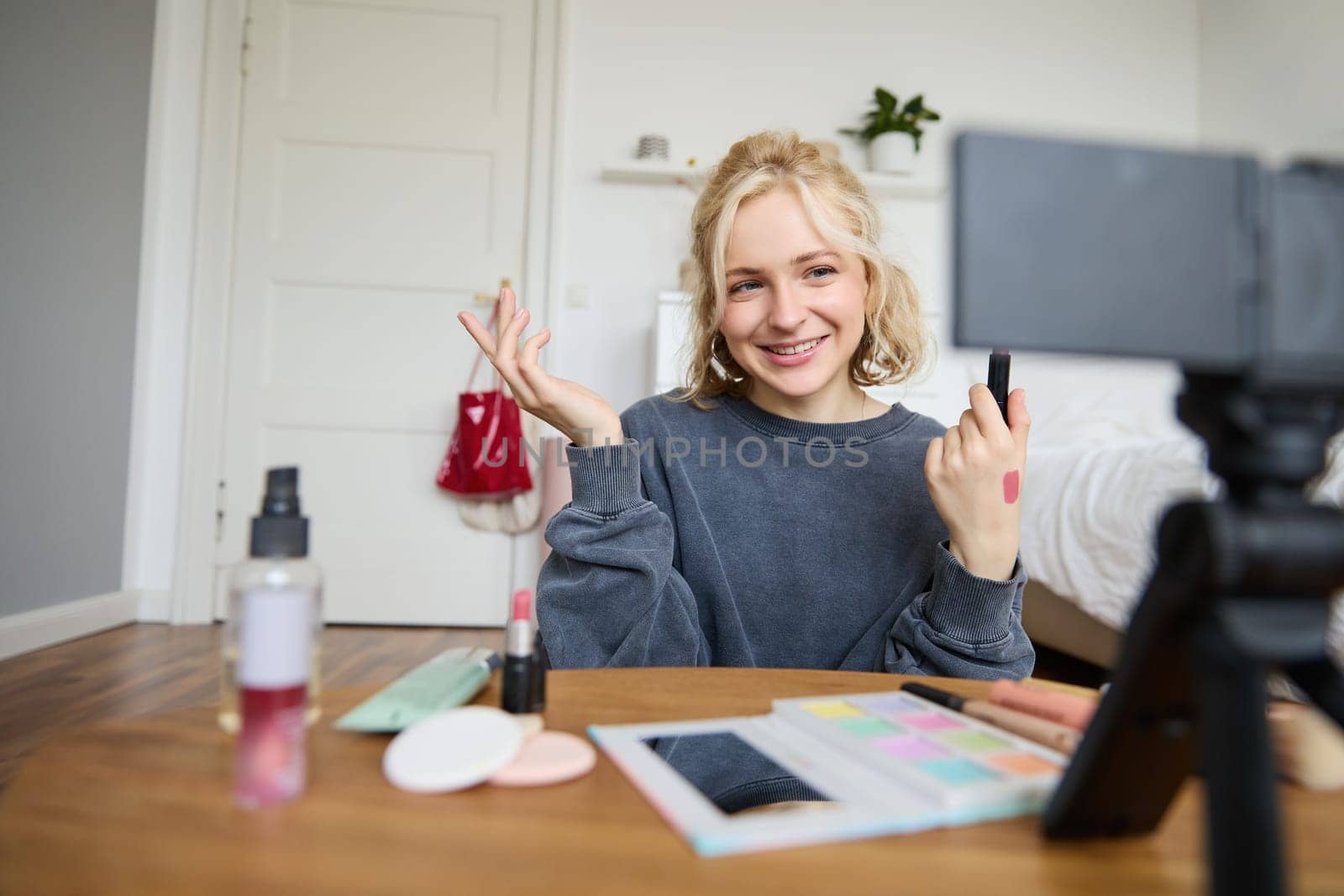 Image of young stylish woman, blogger recording a beauty lifestyle video of her picking best lipstick, showing lip balm swatches on her skin, sitting in front of digital camera in empty room.