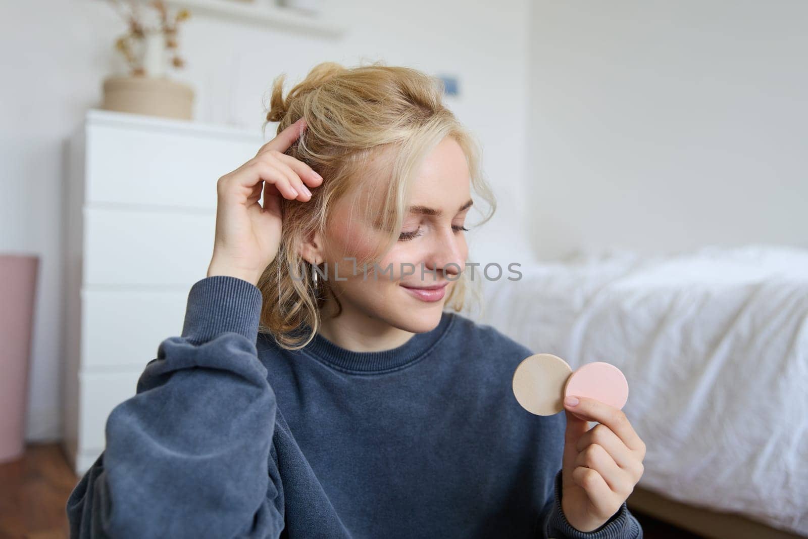 Portrait of young woman chatting on live stream about makeup, sits on floor in bedroom, showing beauty products to followers.