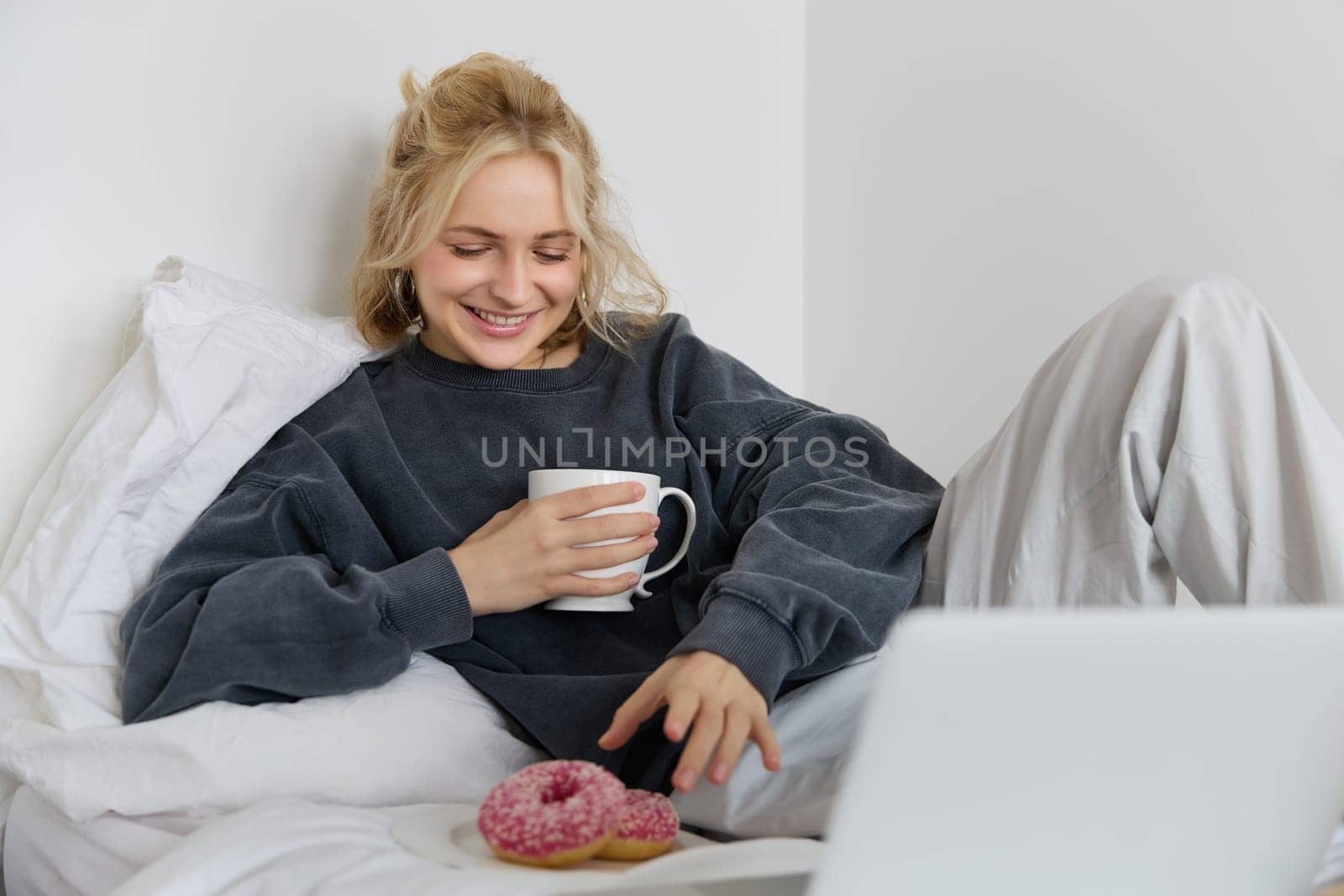 Portrait of happy blond girl, female model lying in bed in cosy clothes, eating and watching movie on laptop, smiling while looking at screen.