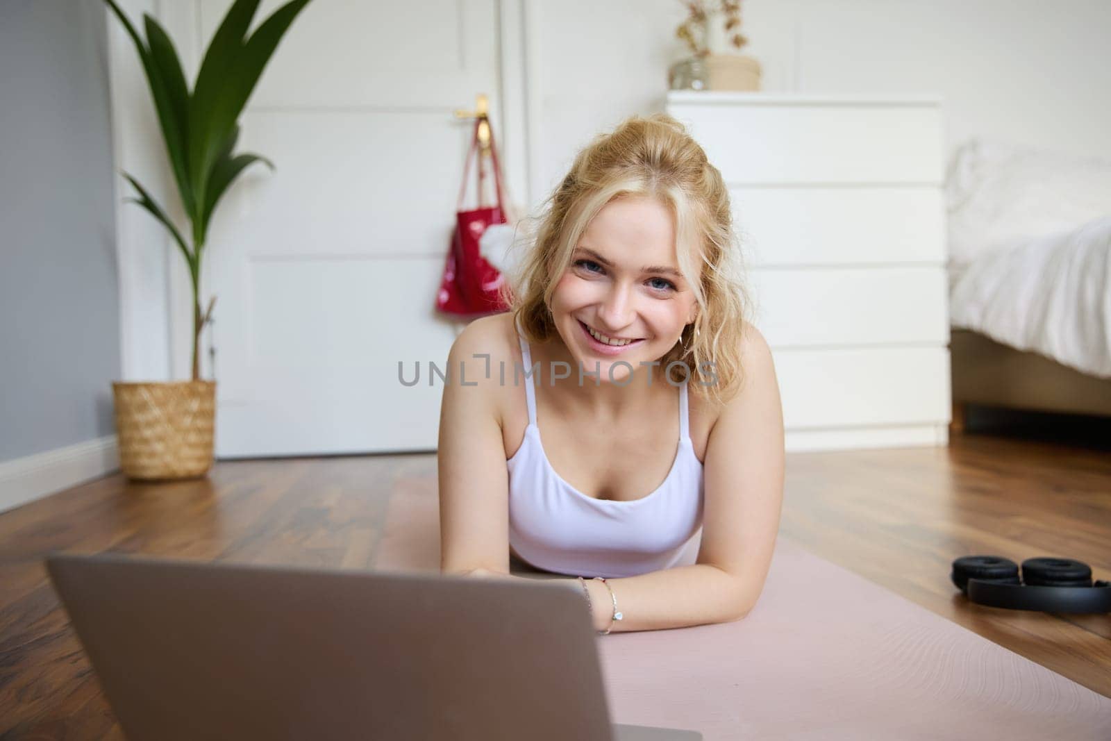 Portrait of beautiful blond woman looking at fitness video tutorials on laptop, lying on rubber yoga mat, following workout instructions online.