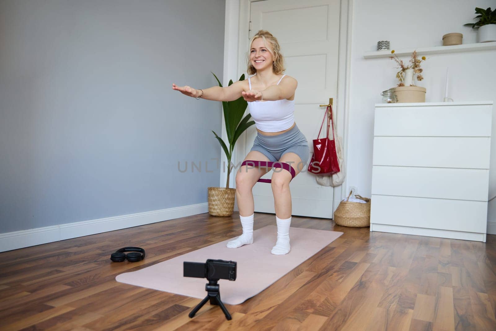 Portrait of woman, fitness instructor at home, recording video about workout, showing how to do leg exercises, squats with elastic resistance band, working out indoors on yoga mat.