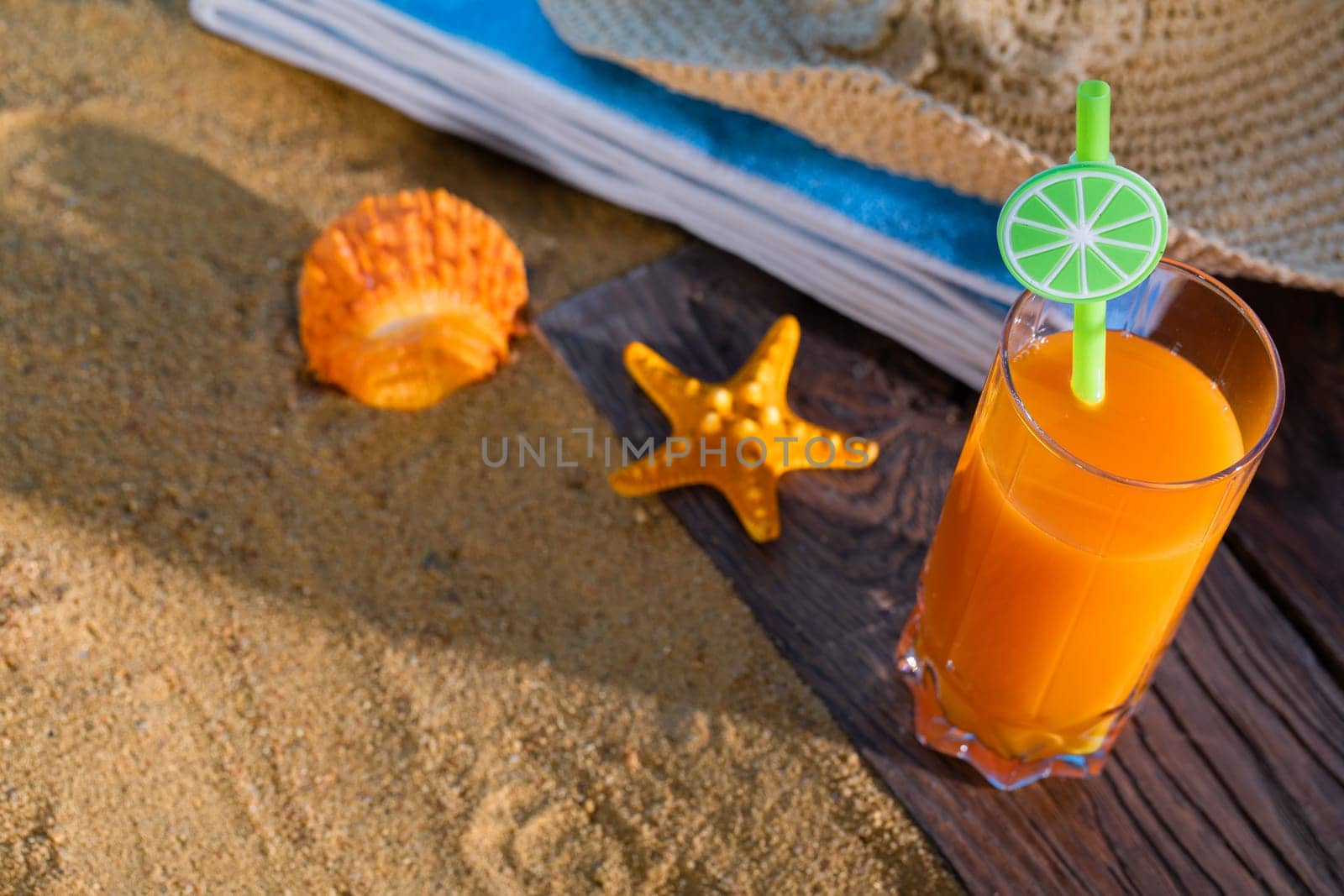 Fresh orange fruit juice stands in a glass with a straw. A beach towel lies on the sandy beach.