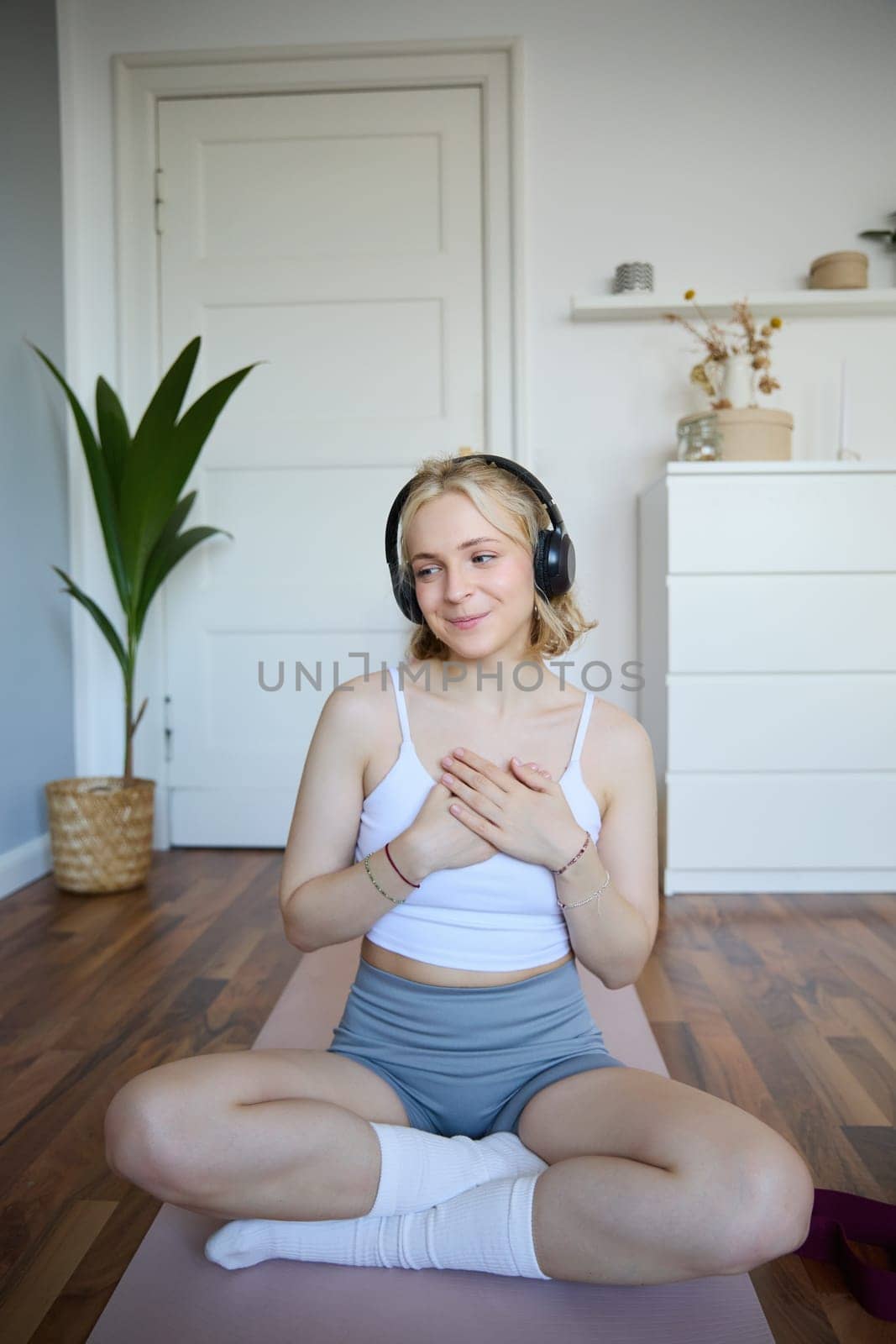 Vertical shot of young sporty woman in headphones, sits on rubber mat after workout, holding hands on chest, looks aside with peaceful, relaxed face expression.