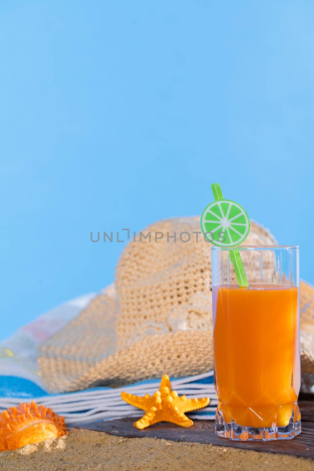 Blue sky. Orange juice in a glass stands on a board covered with sand from the sea beach. Blue beach towel. by fotodrobik