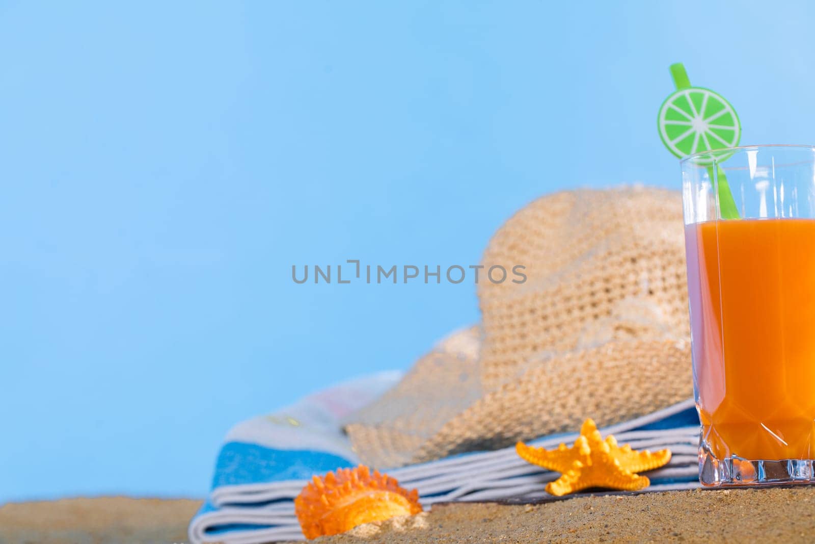 Fresh orange fruit juice stands in a glass with a straw. A beach towel lies on the sandy beach. Blue sky.
