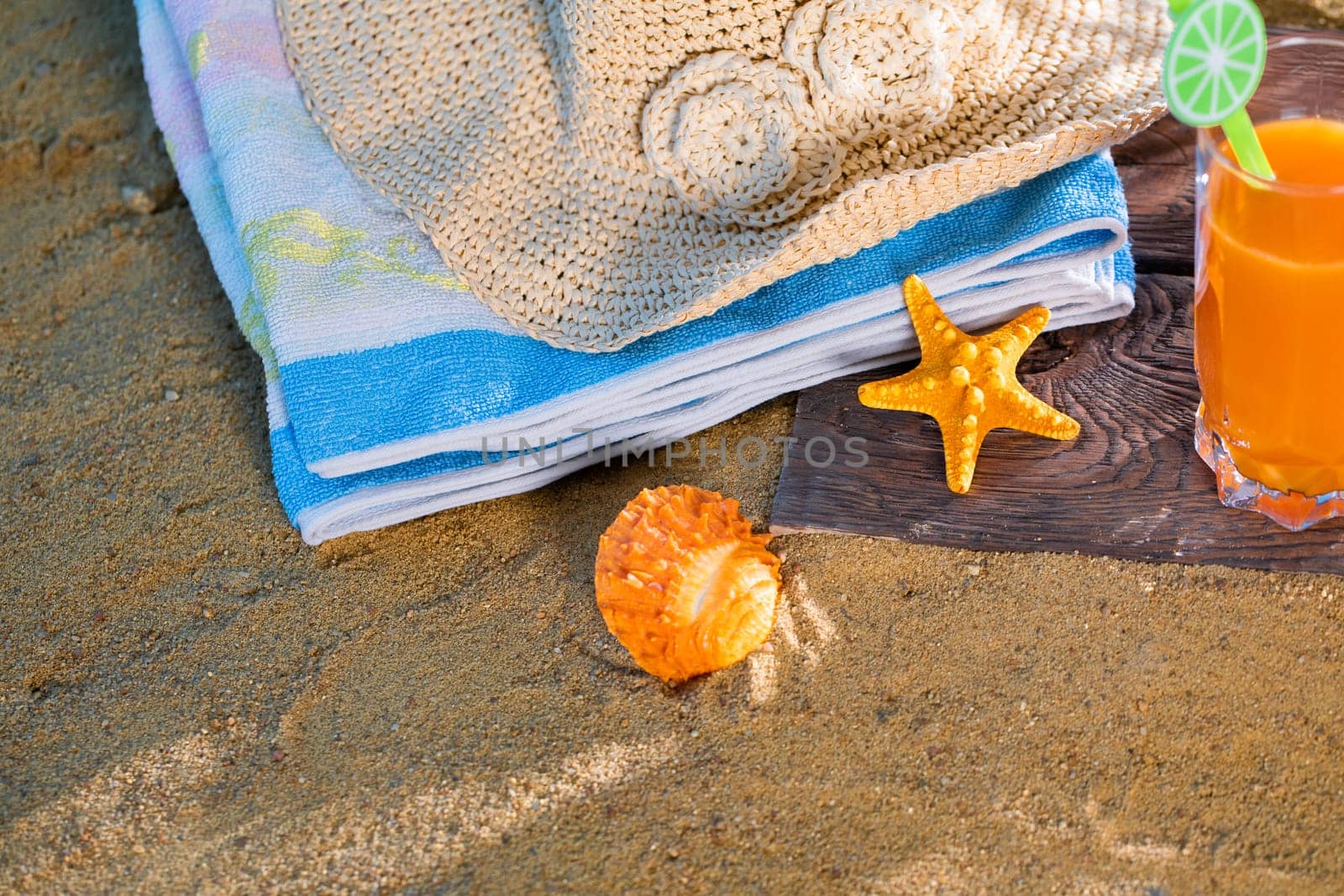 Fresh orange fruit juice stands in a glass with a straw. A beach towel lies on the sandy beach.