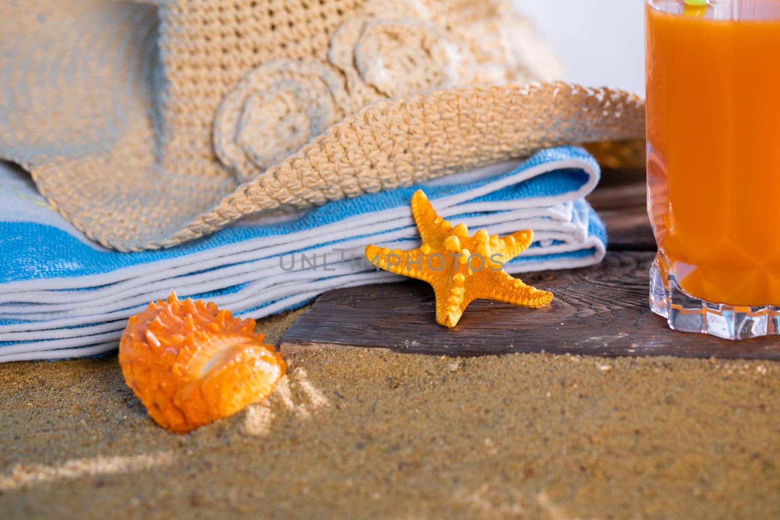 Fresh orange fruit juice stands in a glass with a straw. A beach towel lies on the sandy beach.