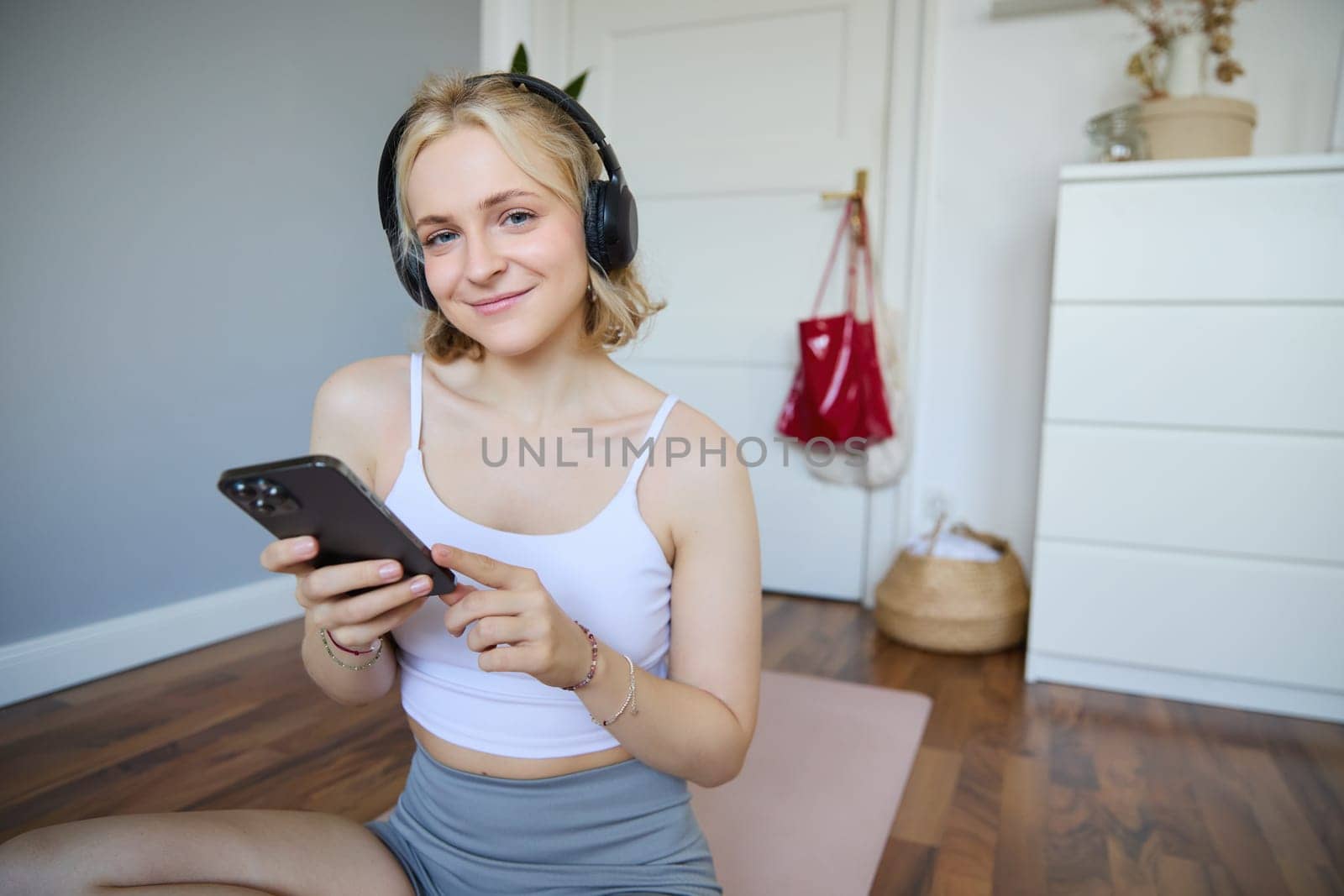 Close up portrait of athletic young woman, holding mobile phone and wearing wireless headphones, workout at home, using sport app during fitness training by Benzoix