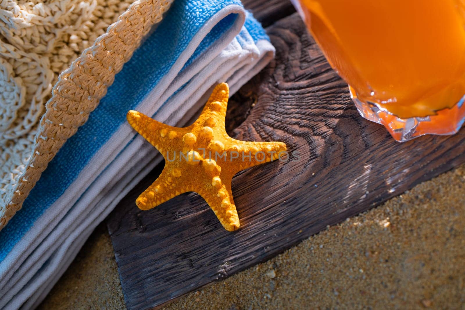 Fresh orange fruit juice stands in a glass with a straw. A beach towel lies on the sandy beach.