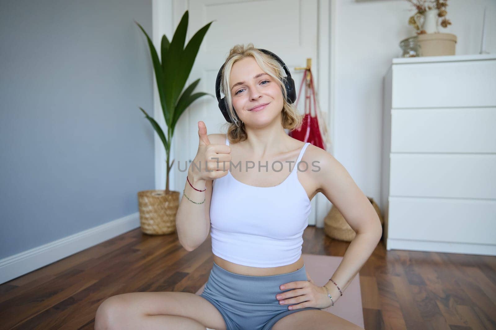 Portrait of beautiful, healthy woman in headphones, showing thumbs up, sitting on rubber mat, recommending yoga online training video, pleased with exercises for home training by Benzoix