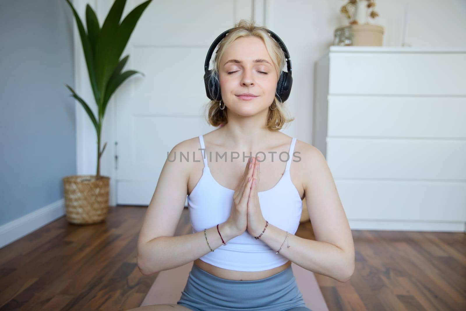 Portrait of young relaxed woman, sits in room in headphones, clasp hands together, meditated, listens to yoga music by Benzoix
