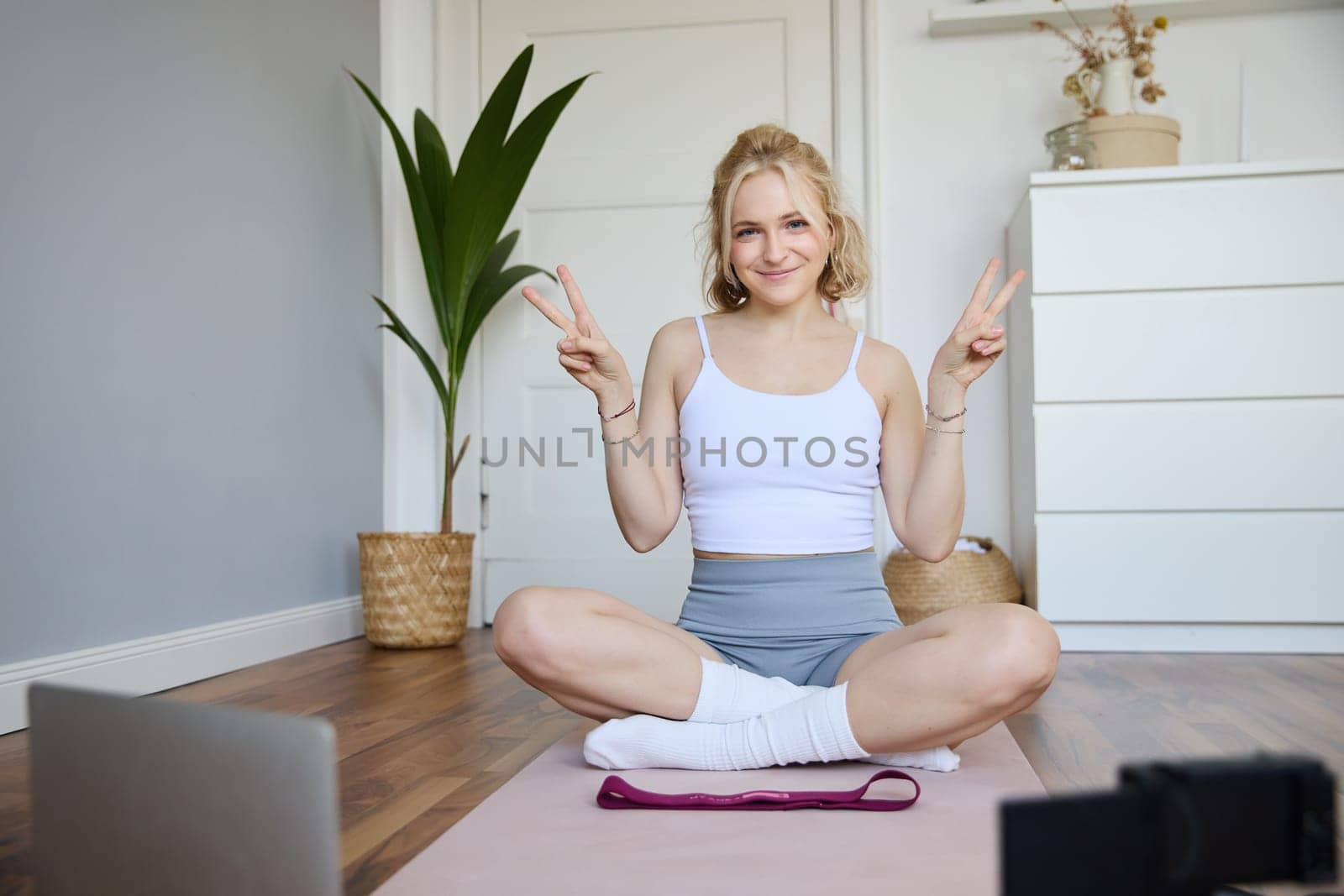 Portrait of cute blond woman shows peace sign, smiles, records yoga vlog on digital camera, workout at home on rubber mat in room.