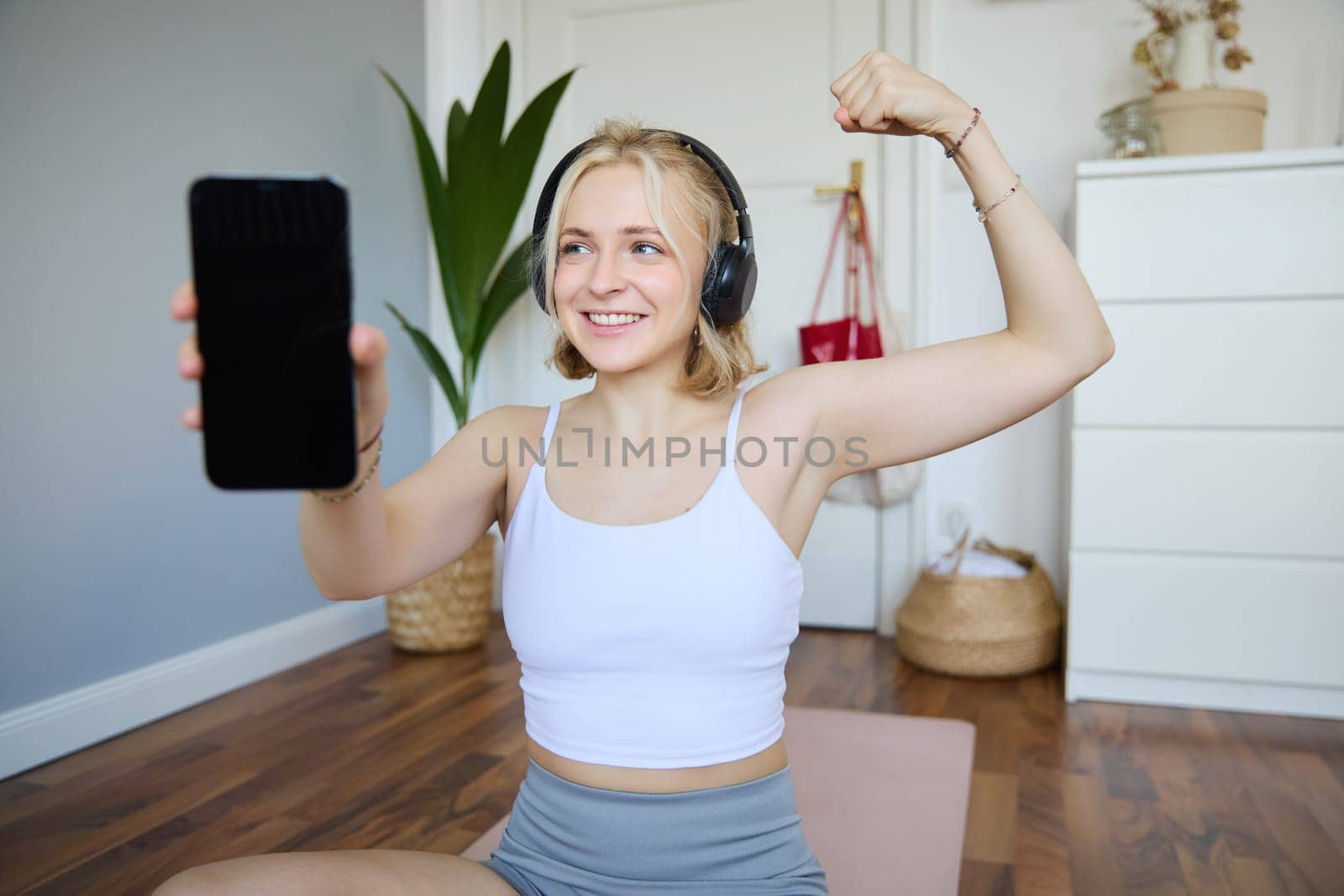Portrait of beautiful and fit young woman, staying healthy working out at home, showing mobile app, blank screen, flexing biceps, recommending smartphone application for exercises by Benzoix