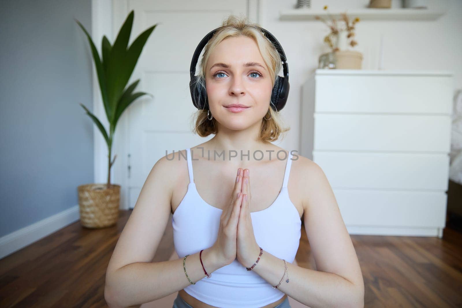 Image of young woman listens to yoga podcast for beginners, holds hands in namaste sign, wears headphones, meditates.