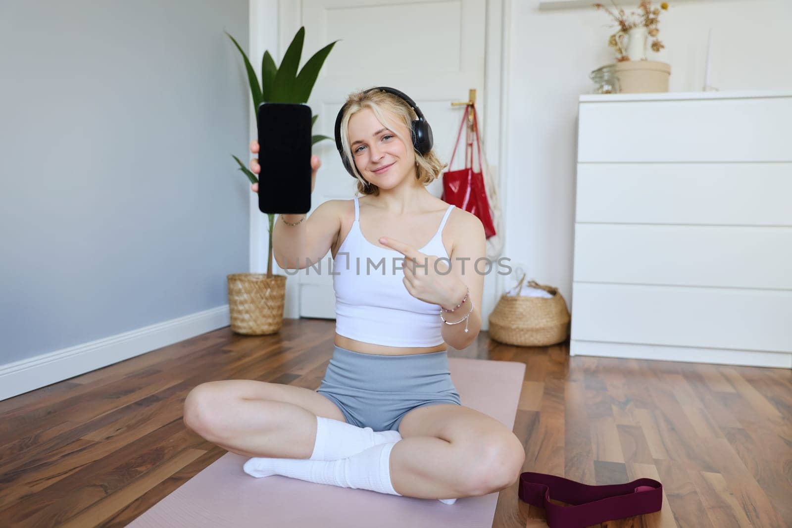 Portrait of fitness instructor showing her favourite workout app, pointing finger at smartphone screen, showing mobile app on camera, smiling and looking satisfied, wearing headphones by Benzoix