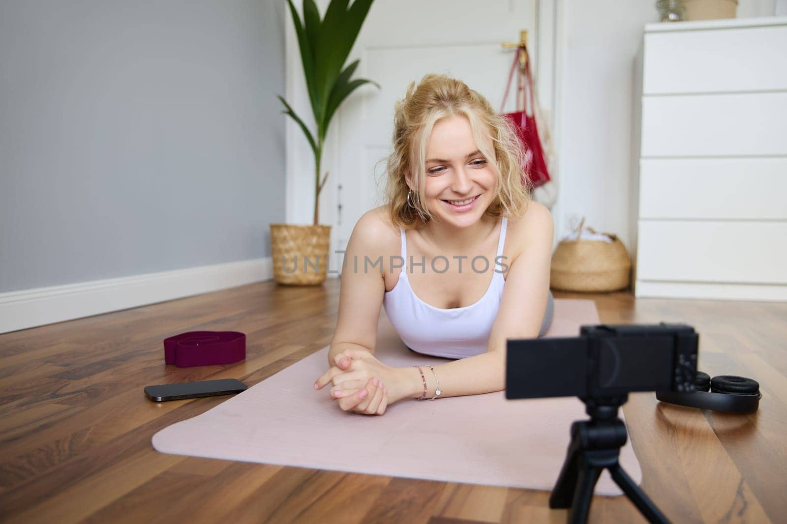 Portrait of young woman, sports vlogger, fitness instructor recording video of herself showing workout exercises, using digital camera, lying on yoga rubber mat.