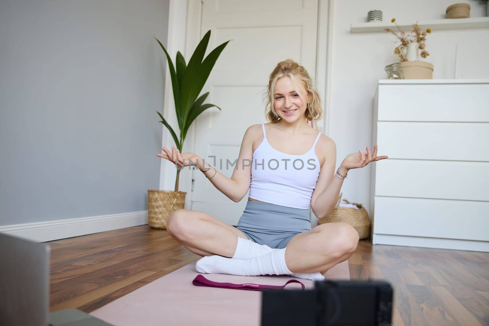Young female athlete, fitness instructor woman sits on floor rubber mat, recording video on digital camera, showing how to workout, explaining exercises.